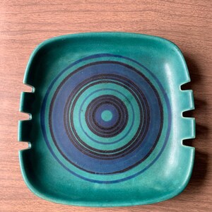 May include: A square, teal ashtray with a concentric circle design in shades of blue and black. The ashtray has four notched rests for cigarettes. The ceramic ashtray is a vintage piece.