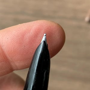 Pu&ograve; includere: Primo piano di un pennino di una penna stilografica nera. Il pennino &egrave; argentato e il corpo della penna &egrave; nero lucido. La penna &egrave; tenuta vicino alla punta di un dito, con uno sfondo sfocato.