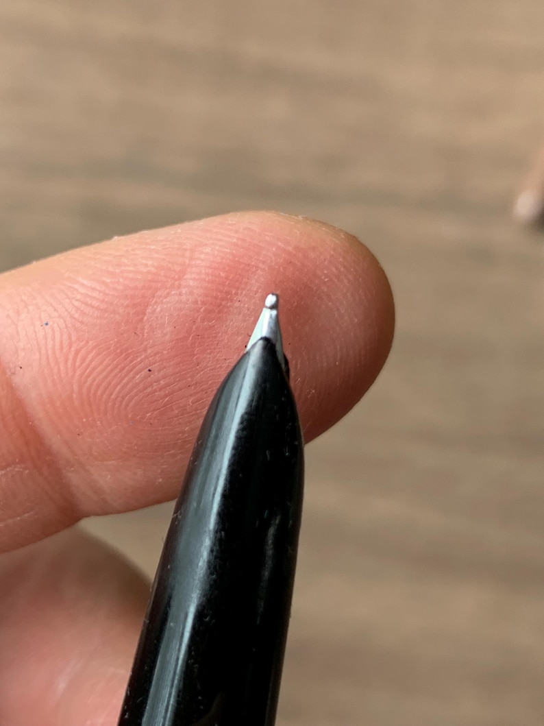 Pu&ograve; includere: Primo piano di un pennino di una penna stilografica nera. Il pennino &egrave; argentato e il corpo della penna &egrave; nero lucido. La penna &egrave; tenuta vicino alla punta di un dito, con uno sfondo sfocato.