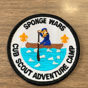 May include: Round patch with the text "SPONGE WARS CUB SCOUT ADVENTURE CAMP". The patch features a cartoon illustration of a person in a blue outfit paddling a canoe on blue water. Two orange fleur-de-lis symbols are on the patch.