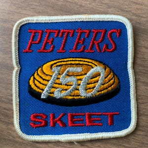 May include: A square embroidered patch with a blue background and a white border. The text PETERS is stitched in red at the top, and SKEET is stitched in red at the bottom. A yellow and black clay target graphic sits in the center with 150 in silver.