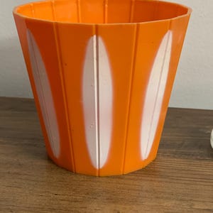 May include: A bright orange plastic plant pot featuring a series of vertical ridges and alternating white oval-shaped accents. The container has a wide, open top and a tapered cylindrical shape with a smooth, uniform finish throughout the exterior.