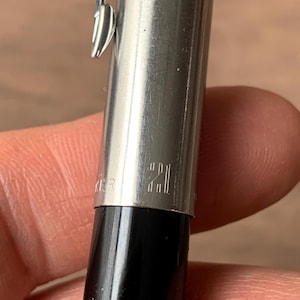 Pu&ograve; includere: Primo piano di una penna argentata e nera. La penna ha un corpo in metallo lucido con il numero "21" inciso. Una clip argentata &egrave; attaccata al corpo della penna. La parte inferiore della penna &egrave; nera.