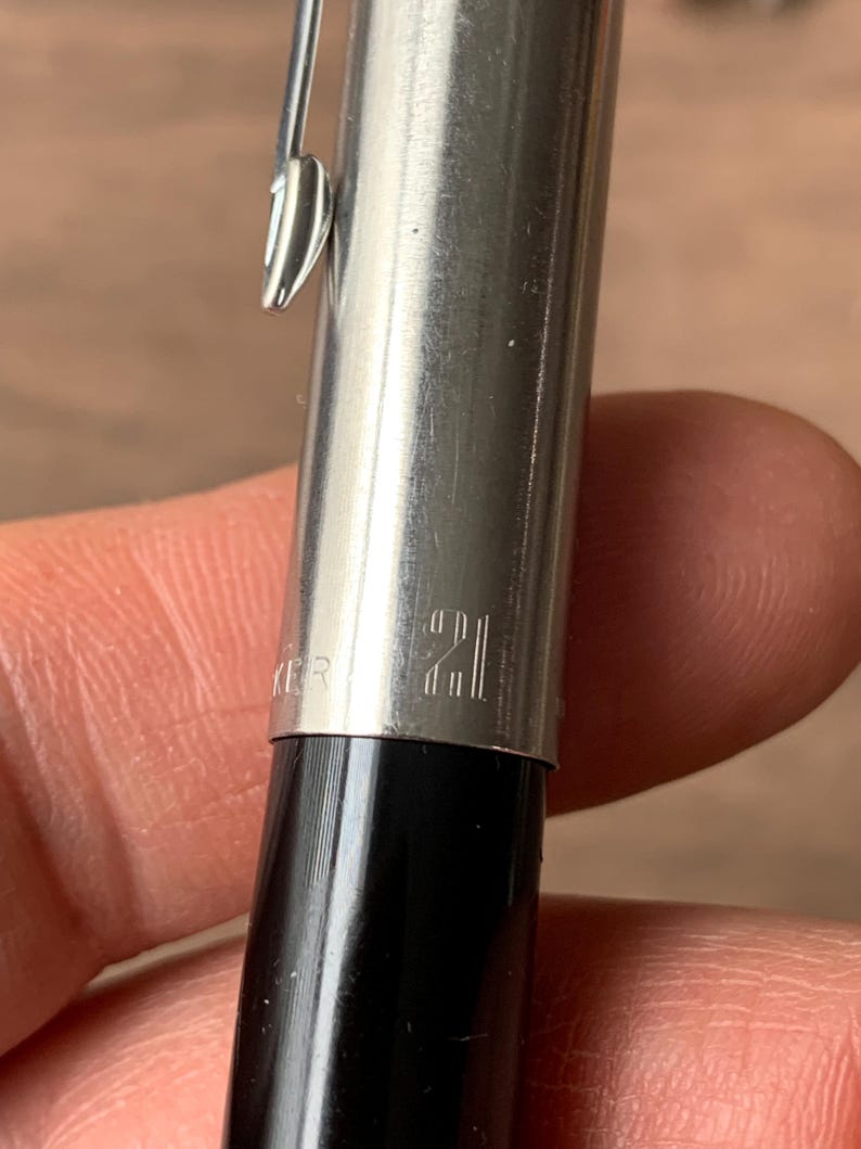 Pu&ograve; includere: Primo piano di una penna argentata e nera. La penna ha un corpo in metallo lucido con il numero "21" inciso. Una clip argentata &egrave; attaccata al corpo della penna. La parte inferiore della penna &egrave; nera.