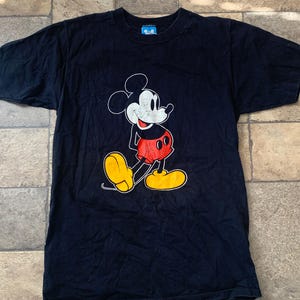 May include: A black t-shirt with a Mickey Mouse design. The cartoon character is in white, red, and yellow. The shirt has a crew neck and short sleeves. The fabric is dark.