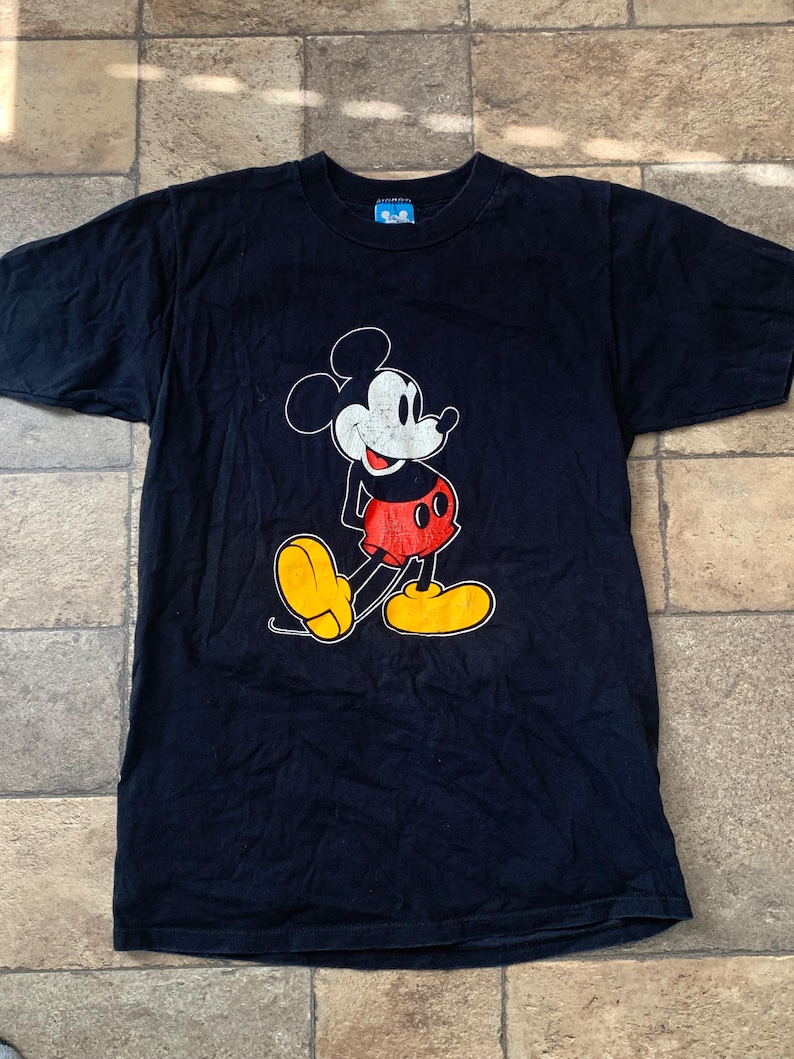 May include: A black t-shirt with a Mickey Mouse design. The cartoon character is in white, red, and yellow. The shirt has a crew neck and short sleeves. The fabric is dark.