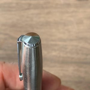 Pu&ograve; includere: Primo piano di una penna d'argento con la parte superiore marrone e una clip d'argento. Il corpo della penna &egrave; cilindrico e ha una finitura metallica lucida. La penna &egrave; tenuta in mano da una persona, con una superficie di legno sullo sfondo.