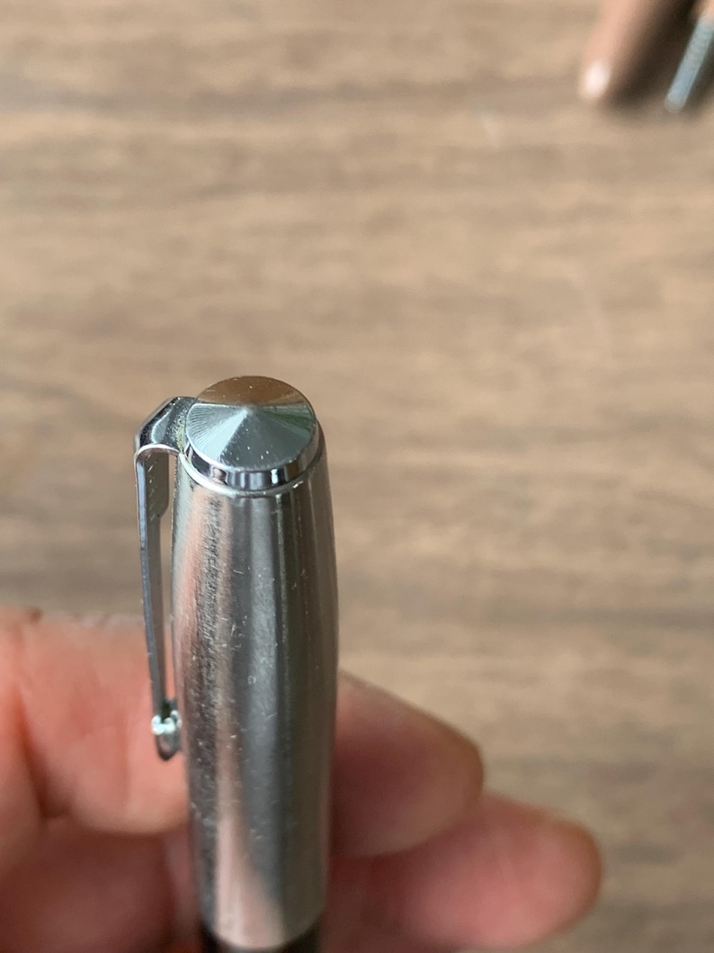 Pu&ograve; includere: Primo piano di una penna d'argento con la parte superiore marrone e una clip d'argento. Il corpo della penna &egrave; cilindrico e ha una finitura metallica lucida. La penna &egrave; tenuta in mano da una persona, con una superficie di legno sullo sfondo.