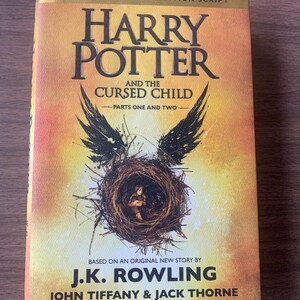 May include: A book titled Harry Potter and the Cursed Child, Parts One and Two, Special Rehearsal Edition Script. The cover features a golden-orange gradient background with a central illustration of a bird's nest containing a small child.
