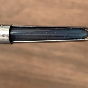 Pu&ograve; includere: Primo piano di un pennino con un serbatoio di inchiostro blu scuro. Il pennino &egrave; racchiuso in un alloggiamento di metallo argentato. Lo sfondo &egrave; una superficie di legno sfocata.