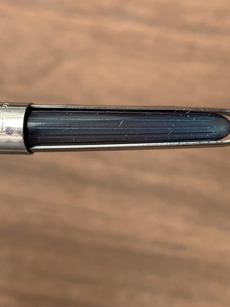 Pu&ograve; includere: Primo piano di un pennino con un serbatoio di inchiostro blu scuro. Il pennino &egrave; racchiuso in un alloggiamento di metallo argentato. Lo sfondo &egrave; una superficie di legno sfocata.