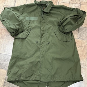 May include: An olive green field jacket featuring a high collar, a front zipper closure with a storm flap, two large flap pockets at the waist, and a drawstring waist. The jacket has long sleeves with adjustable cuffs and a fishtail hem.