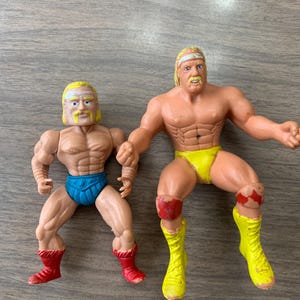 May include: Two plastic action figures of muscular wrestlers. One figure wears blue trunks and red boots, the other wears yellow trunks and boots. Both have blonde hair and mustaches.