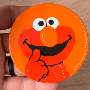 May include: A round, orange button featuring a smiling cartoon character with red fur, a large orange nose, and white eyes. The character is giving a thumbs up. The text "© Jim Henson Productions" is printed on the right side.
