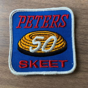 May include: A square embroidered patch with a blue background and a white border. It features the red text PETERS at the top and SKEET at the bottom. In the center is a yellow clay target graphic with the white embroidered number 50 overlaid on it.