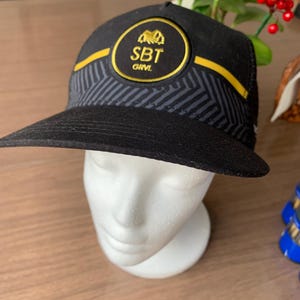 May include: Black trucker hat with a curved brim. The hat features a circular patch with gold lettering and a logo. The brim has a geometric pattern. The text "SBT GRVL" is visible on the patch.