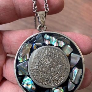May include: A circular pendant necklace featuring a central silver-toned disc with an embossed sun calendar design, surrounded by a border of iridescent abalone shell fragments set in black, suspended from a silver-toned twisted chain.