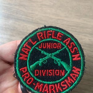 May include: Round black patch with red and green embroidered text and design. The text reads "NATL RIFLE ASSN JUNIOR DIVISION PRO-MARKSMAN". Two green rifles are crossed in the center of the patch.