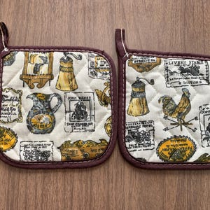 May include: Two quilted pot holders with a cream-colored background and a repeating vintage-style print featuring illustrations of a rooster, coffee grinders, and other antique motifs. The edges are trimmed with a dark brown fabric.