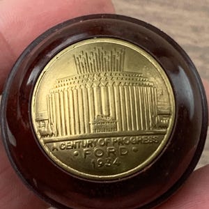 May include: A circular brown knob featuring a gold-colored metal inlay. The center depicts a building with vertical columns and the text A CENTURY OF PROGRESS FORD 1934. The object has a smooth, polished finish and a rounded, domed profile.