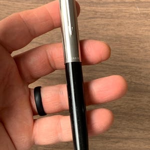 Pu&ograve; includere: Una penna nera e argento con finitura lucida. La penna ha un cappuccio argentato e un fusto nero. La penna &egrave; tenuta in mano. La penna ha la scritta "PARKER" stampata sul cappuccio argentato.
