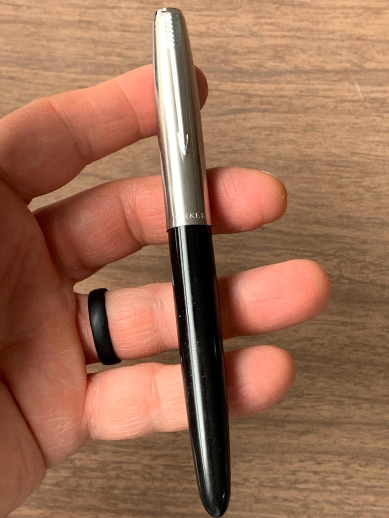 Pu&ograve; includere: Una penna nera e argento con finitura lucida. La penna ha un cappuccio argentato e un fusto nero. La penna &egrave; tenuta in mano. La penna ha la scritta "PARKER" stampata sul cappuccio argentato.