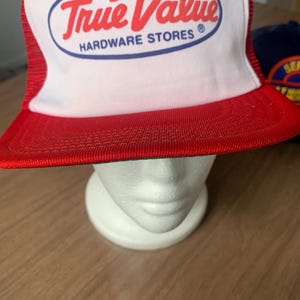 May include: A red and white trucker hat with the text "True Value HARDWARE STORES" in red script within a blue oval. The hat has a red brim and a white front panel.