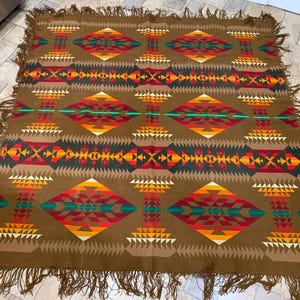 May include: A brown rectangular throw blanket featuring a repeating geometric pattern of diamonds and triangles in shades of red, orange, yellow, and teal. The blanket is finished with long, knotted brown fringe along all four edges.