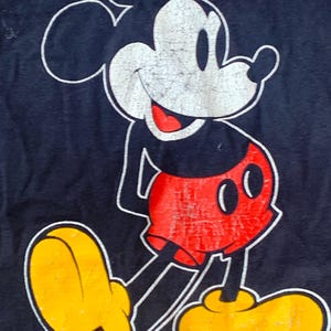 May include: A navy blue t-shirt with a distressed Mickey Mouse graphic. The cartoon mouse has a white face, red shorts, yellow shoes, and a black body. The outline is white.