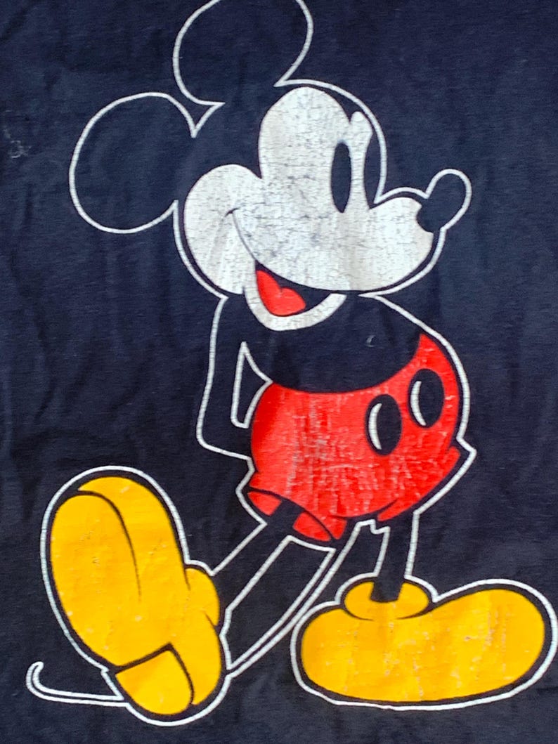 May include: A navy blue t-shirt with a distressed Mickey Mouse graphic. The cartoon mouse has a white face, red shorts, yellow shoes, and a black body. The outline is white.