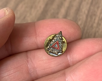 Op de afbeelding: Kleine, ronde, goudkleurige speld met een driehoekig ontwerp. De driehoek heeft een rood midden en is omgeven door een zilverkleurige, getextureerde rand. De speld wordt in de hand van een persoon gehouden.