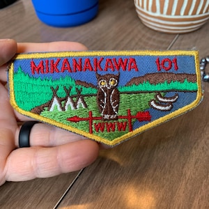 May include: A blue and yellow embroidered patch with the text "Mikanakawa 101". The patch features a brown owl with yellow eyes, a red arrow, and a green forest scene.