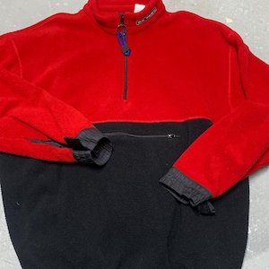 May include: A red and black fleece pullover with a half-zip closure. The top half is red, and the bottom half is black. The sleeves have black cuffs. The collar has a small label with white text. A blue and black pull is attached to the zipper.