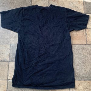 May include: A dark navy blue t-shirt with short sleeves, lying flat on a tiled surface. The shirt appears to be made of a soft fabric and has a simple, classic design. The fabric has visible wrinkles.