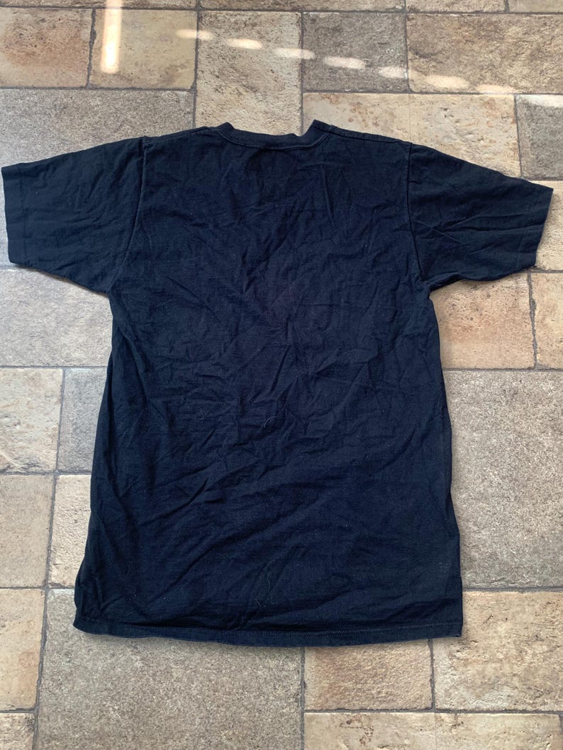 May include: A dark navy blue t-shirt with short sleeves, lying flat on a tiled surface. The shirt appears to be made of a soft fabric and has a simple, classic design. The fabric has visible wrinkles.