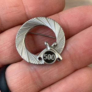 May include: Silver-toned bowling pin brooch with a circular design. The brooch features a ring with a textured pattern and a small emblem with bowling pins and the number 500. The pin is held in a hand.