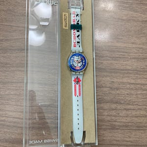 May include: A Swatch wristwatch in a clear plastic case. The white strap features Cyrillic text, a rocket graphic, and a blue watch face with a dog illustration and the year 1964. The watch includes a dark green keeper and a white buckle.