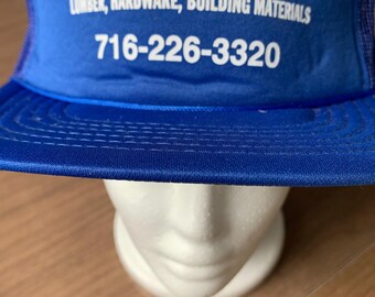 May include: A blue trucker hat with white text that reads "AVON LUMBER INC." and "LUMBER, HARDWARE, BUILDING MATERIALS." The hat also displays a phone number. The hat is displayed on a white mannequin head.