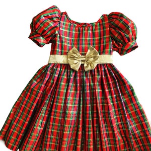 May include: A red and green plaid dress with a gold bow and puff sleeves. The dress has a festive holiday theme.