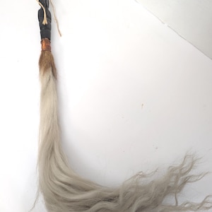 Puede incluir: Un objeto decorativo con un mango de madera negra tallada, una banda de cobre y una larga cola de pelo gris claro. El mango está adornado con una pequeña figura tallada y atado con hilo.