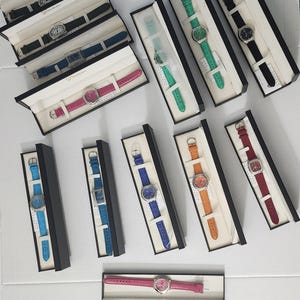 Pode incluir: Uma coleção de doze relógios de pulso, cada um apresentado numa caixa retangular preta. Os relógios possuem braceletes de couro texturizado em várias cores, incluindo preto, azul, verde, rosa, laranja e vermelho, com caixas de metal prateado.