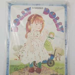 May include: A clear plastic carrying case containing a book titled Dolly Dolly Barbara. The cover features an illustration of a girl with reddish-brown hair in a white floral dress, holding a small doll in a blue cart, with flowers and trees.