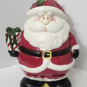 Op de afbeelding: Een keramieken kerstmanfiguur in een rood pak met witte randen, een zwarte riem met een gele gesp en zwarte laarzen. Het figuur draagt een rode muts met hulstblaadjes en houdt een wit cadeau vast met rode strepen en een groene strik.