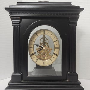 May include: A black mantel clock with a rectangular frame and fluted columns. The clock face is circular with Roman numerals and a visible gold-toned gear mechanism behind the hands. The clock is encased in glass and stands on four small feet.