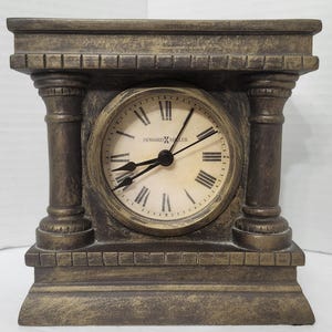 May include: A Howard Miller mantel clock with a dark bronze-toned finish and a rectangular frame featuring two side columns. The circular clock face has a cream background, black Roman numerals, and black hands, with the brand name printed in the centre.