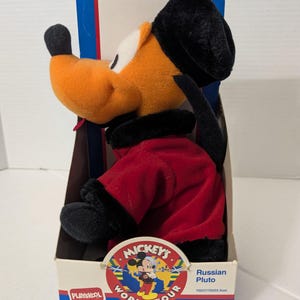 Pode incluir: Um brinquedo de pelúcia do personagem Pluto da coleção Mickey's World Tour, vestindo um casaco vermelho com acabamento preto e um chapéu de pele preto. O brinquedo está na embalagem original de papelão com o texto Playskool e Russian Pluto.