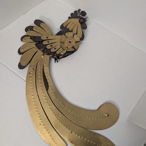 May include: A wall-mounted bird sculpture crafted from layered brass and dark metal components. The bird features a prominent crest, a circular chest detail, and long, sweeping tail feathers adorned with small, raised metal studs.