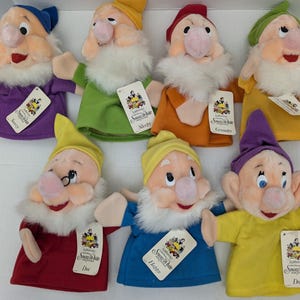 May include: A set of seven plush hand puppets representing the seven dwarfs from Snow White. Each puppet features a soft white beard, a colourful hat, and a matching tunic. Each puppet includes a white paper tag with the Snow White logo.