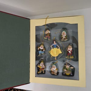 Op de afbeelding: Een set van acht Sneeuwwitje en de zeven dwergen ornamenten in een crèmekleurige doos. De collectie bevat een Sneeuwwitje-figuur met een gele rok en blauw lijfje, vergezeld door zeven verschillende dwergfiguren in individuele vakjes.