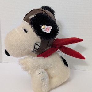 Könnte beinhalten: Ein Snoopy-Plüschtier mit brauner Fliegermütze mit schwarzem Besatz und rotem Schal. Der weiß-schwarze Hund hat ein gesticktes Lächeln und ein Steiff-Etikett.